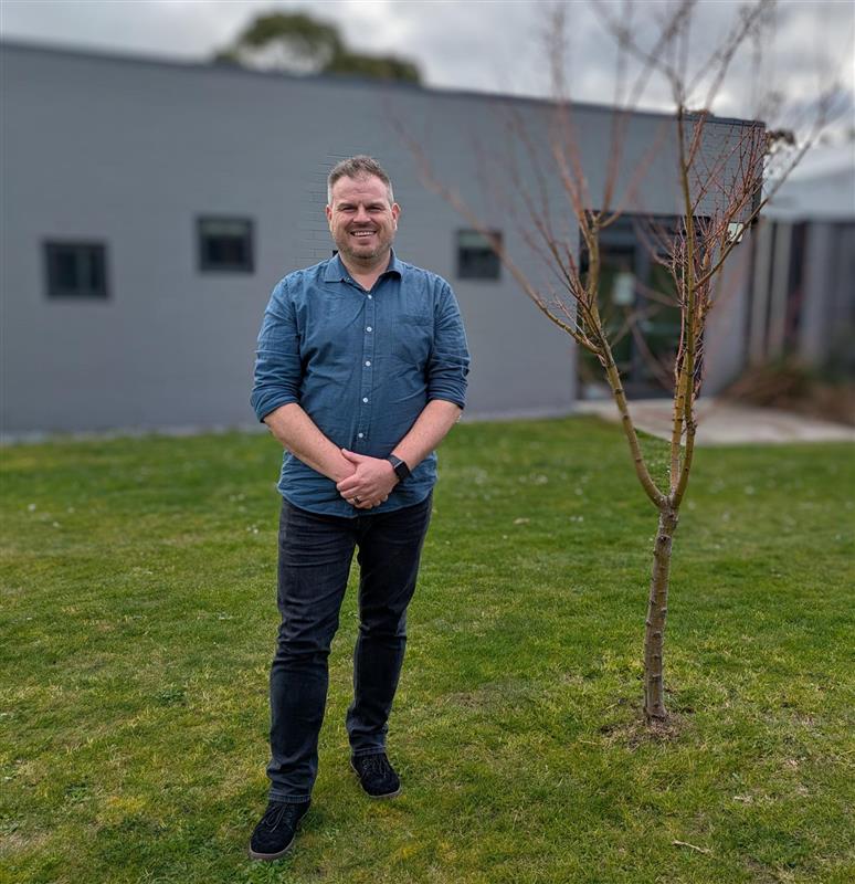 Man standing next to a bare tree on a cloudly day, the building in the background is grey and the grass is green.
